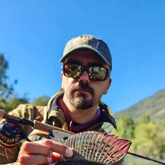 A grayling's tall red-spotted dorsal fin held up to the camera, with Aleksandar Gasevic behind it under a blue sky.